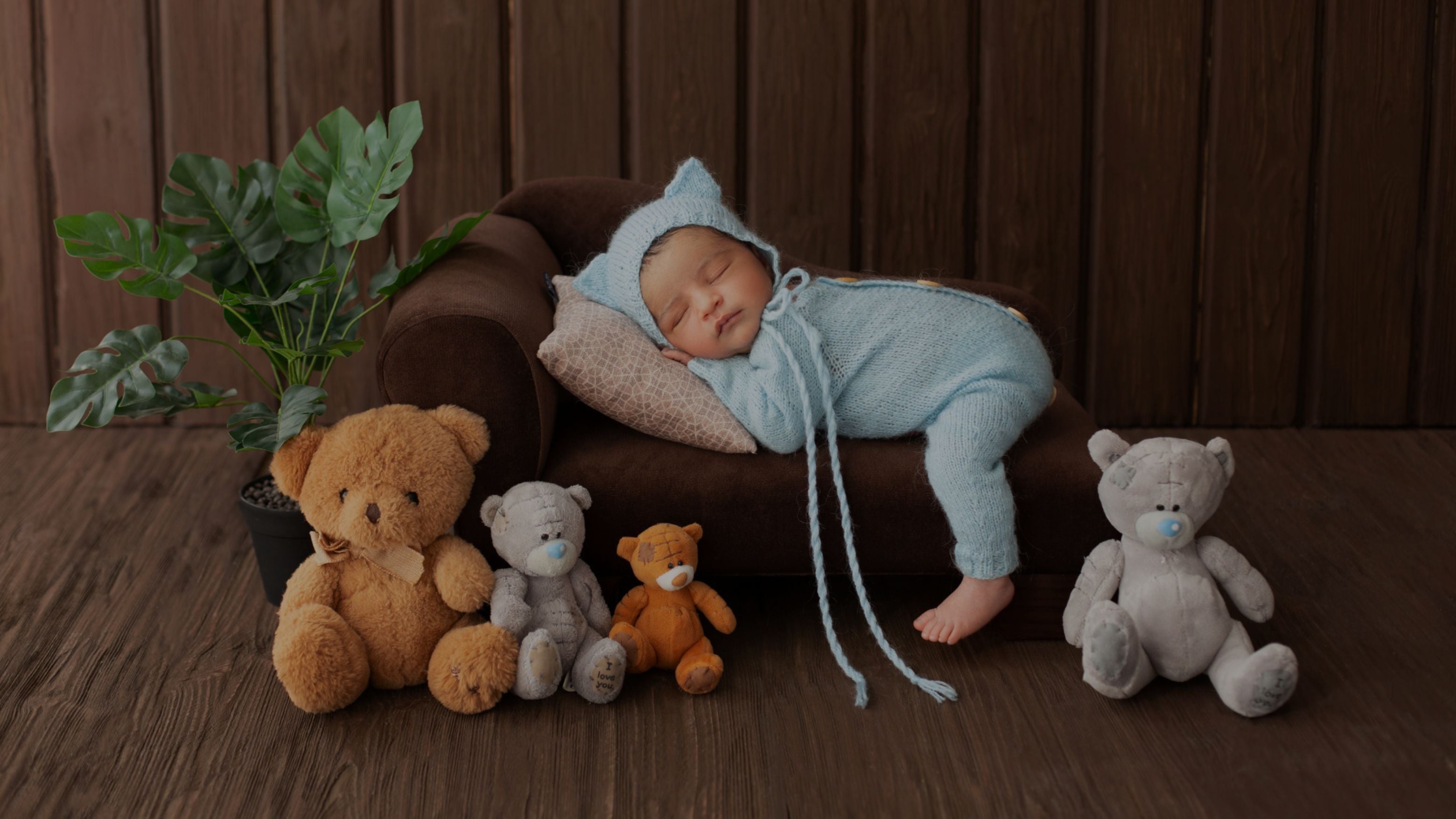 Baby in a blue outfit lying on a brown couch with teddy bears around