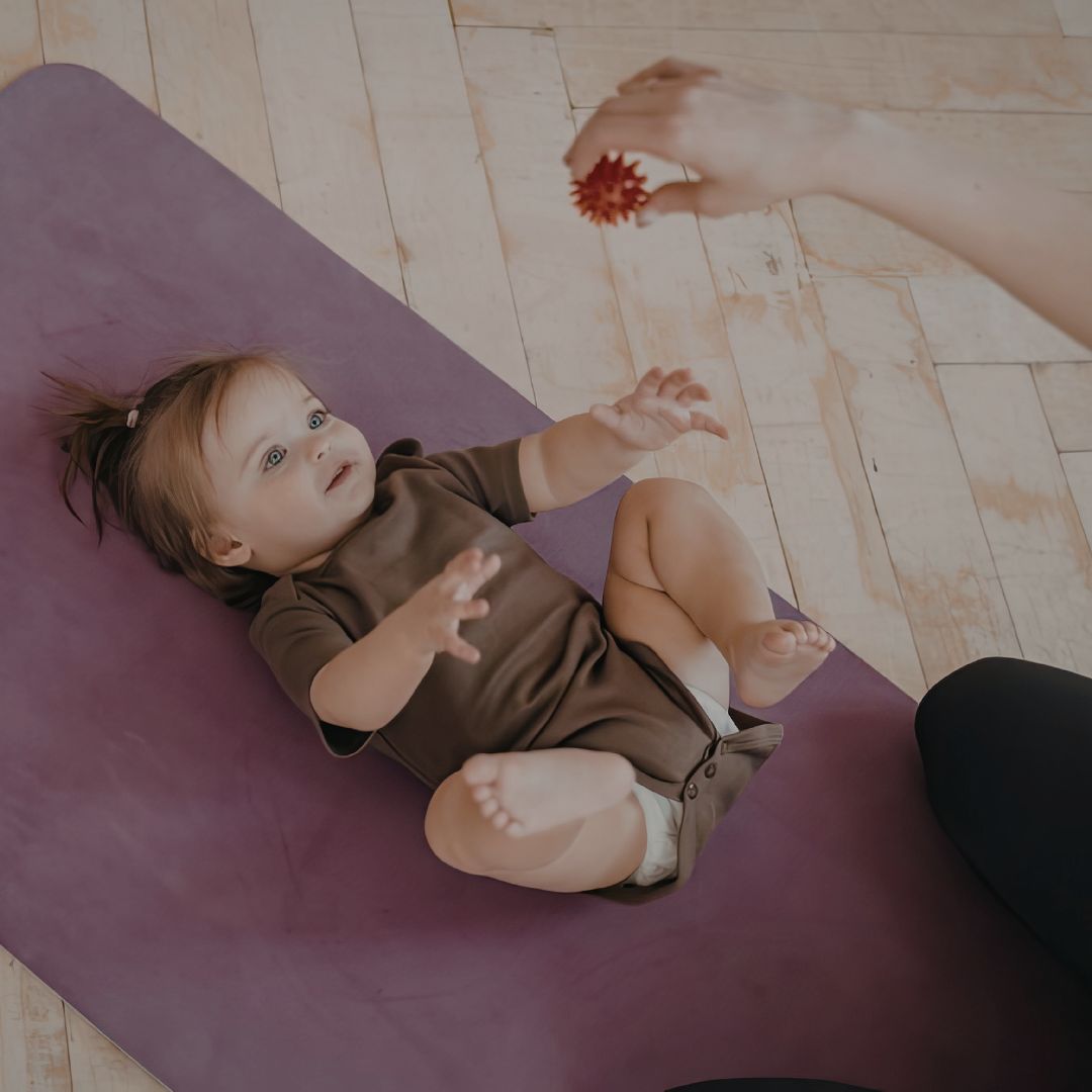 Child lying on a purple mat with a person's hand holding a red object nearby.